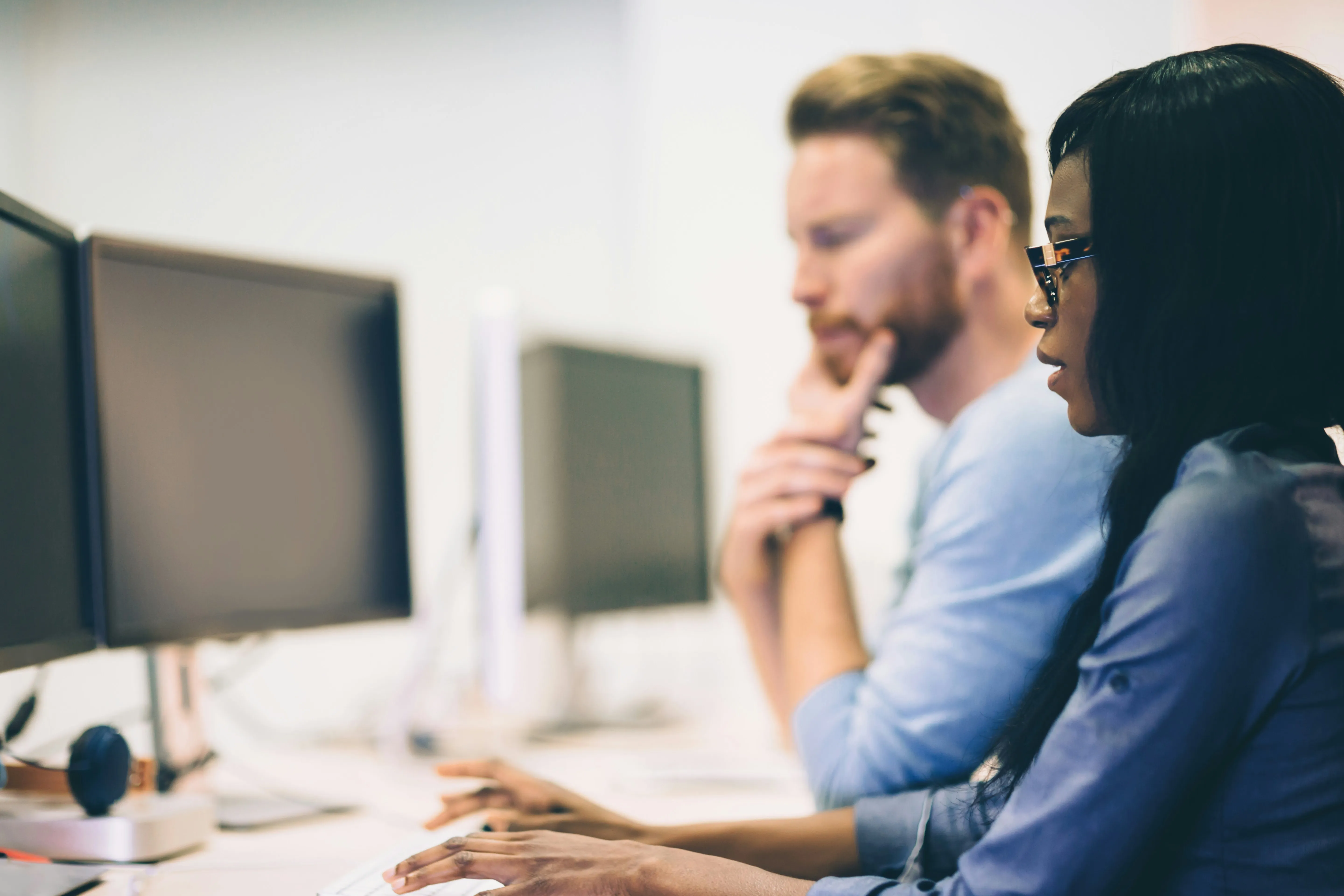 Two coworkers focused on computer screens, a woman typing and a man thinking with his hand on his chin.