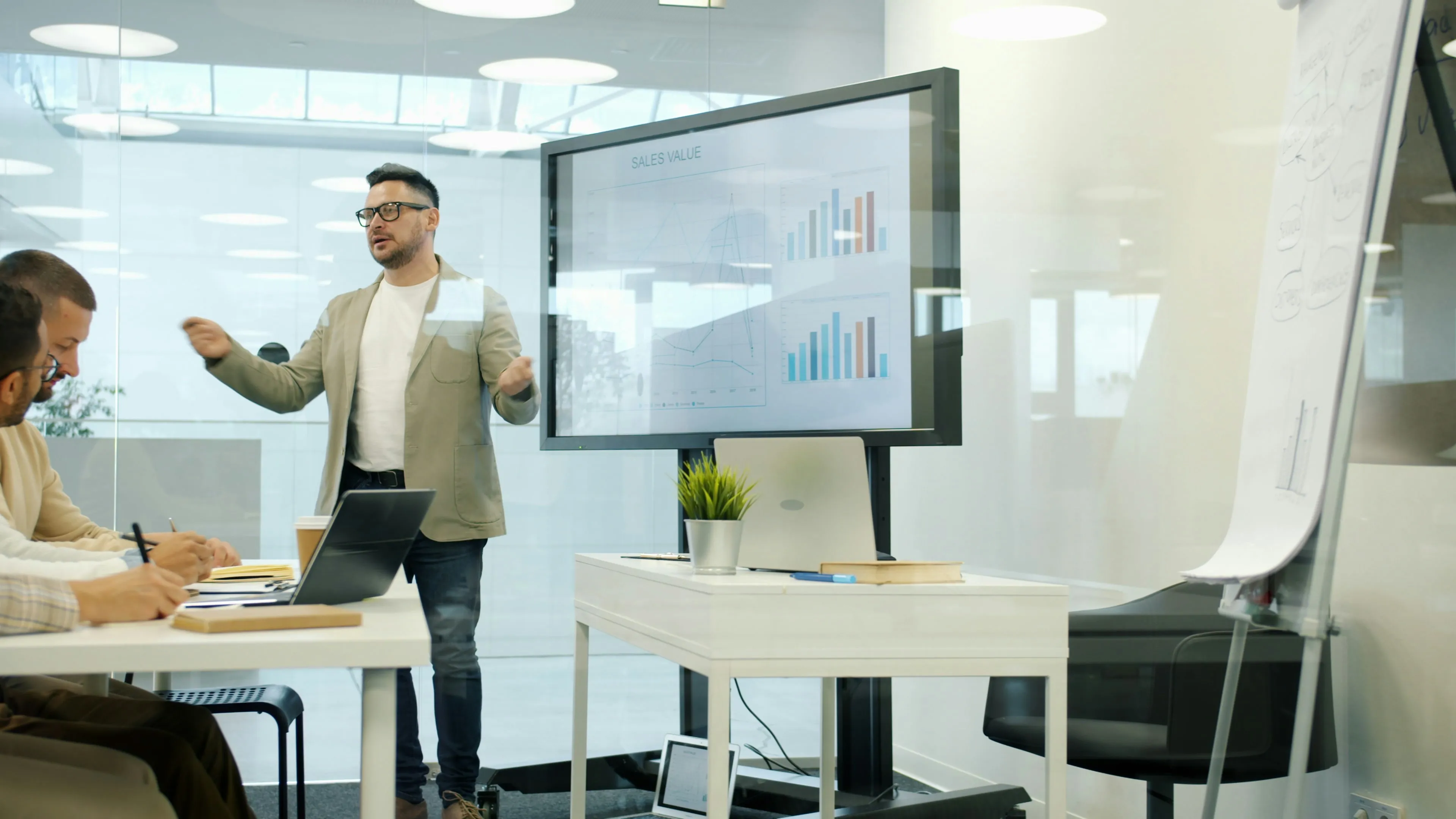 Man in glasses and beige jacket presenting sales value charts on a screen to colleagues in a modern office.