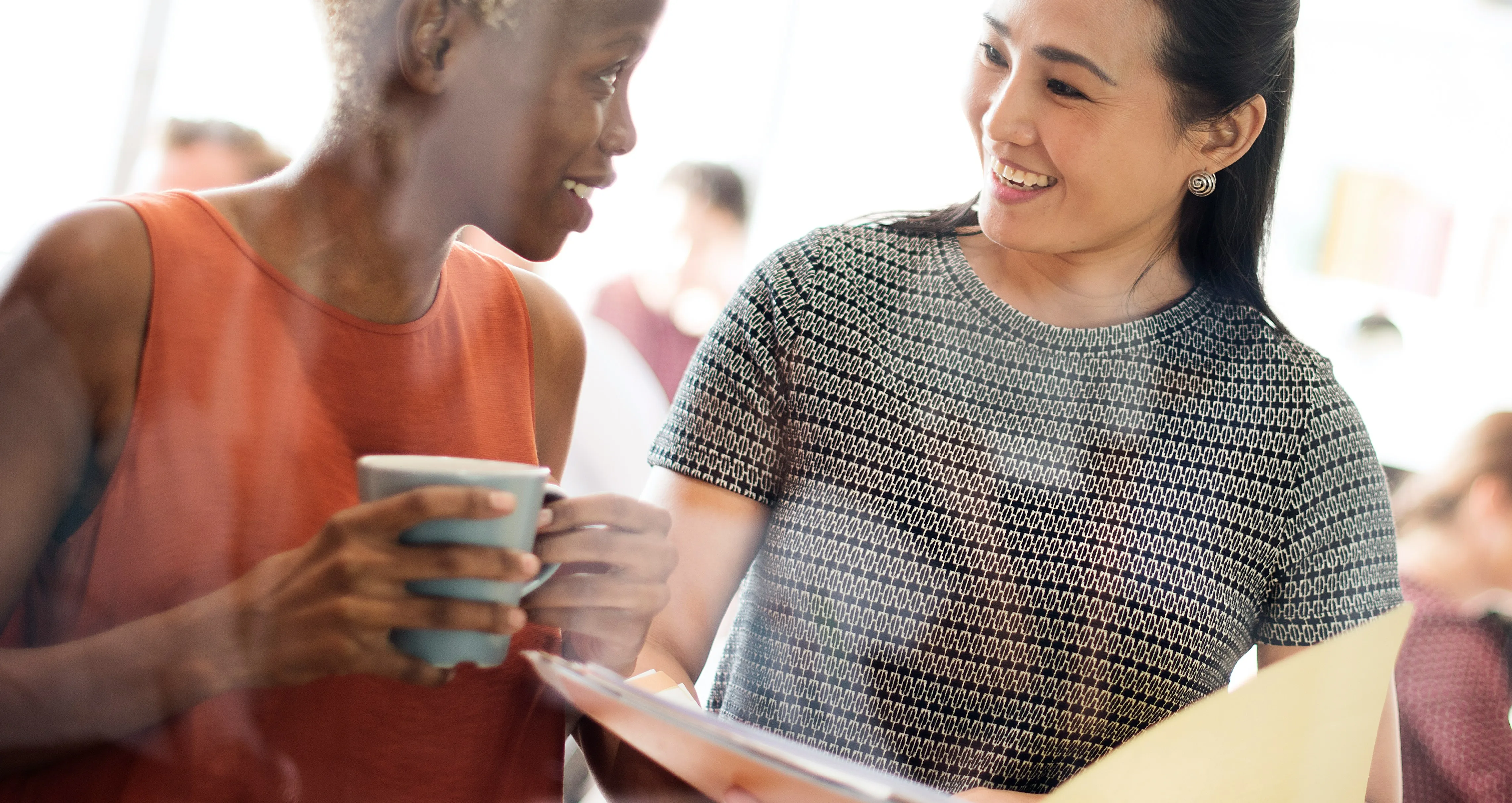 Two women smiling and talking, one holding a mug and the other holding an open folder with papers.