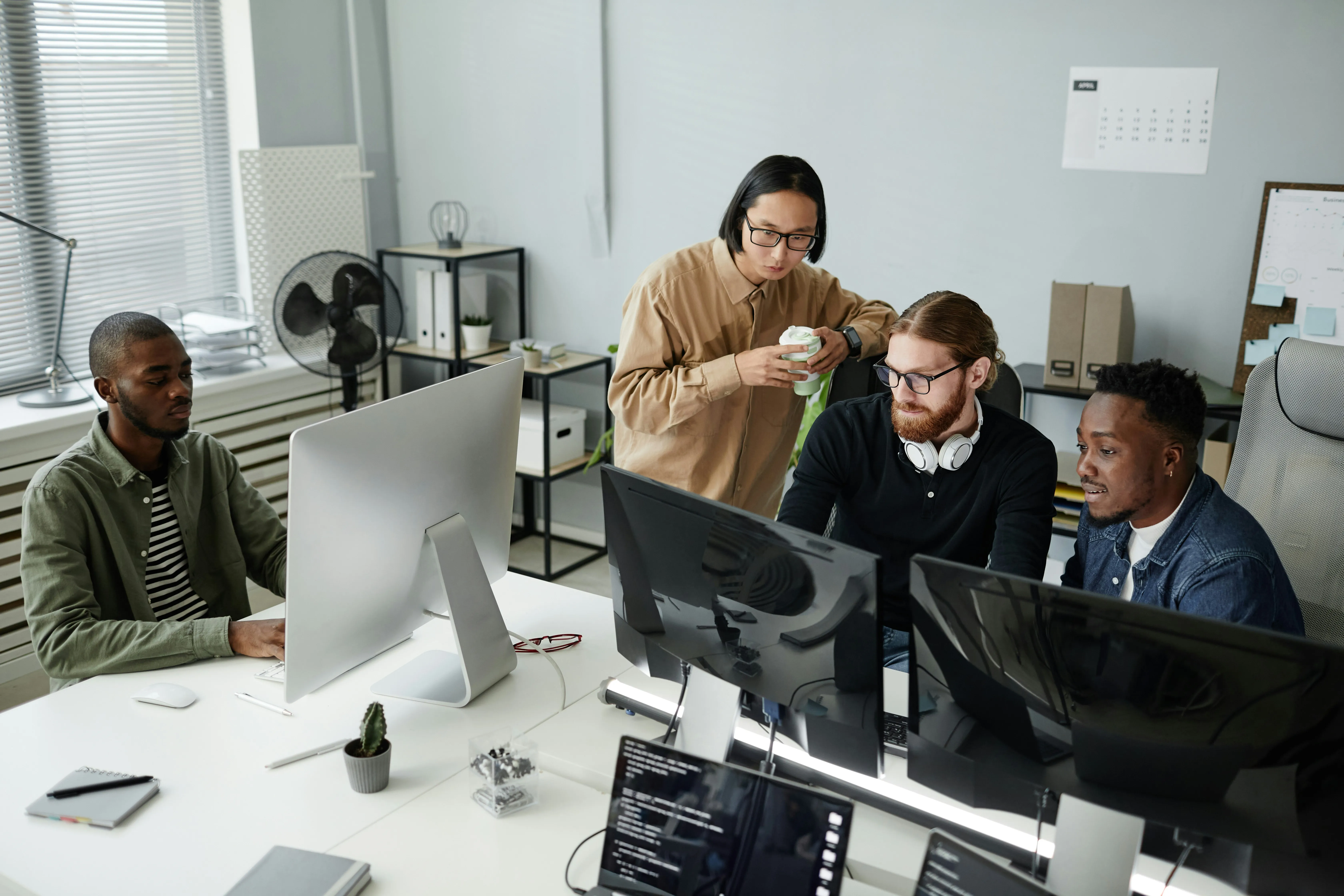 Four diverse coworkers at a modern office desk with multiple computer monitors, collaborating and working together.