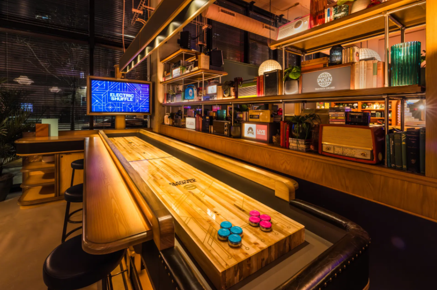 Cozy indoor game area featuring a wooden shuffleboard table with blue and pink pucks, surrounded by bar stools and shelves with vintage radios and books.