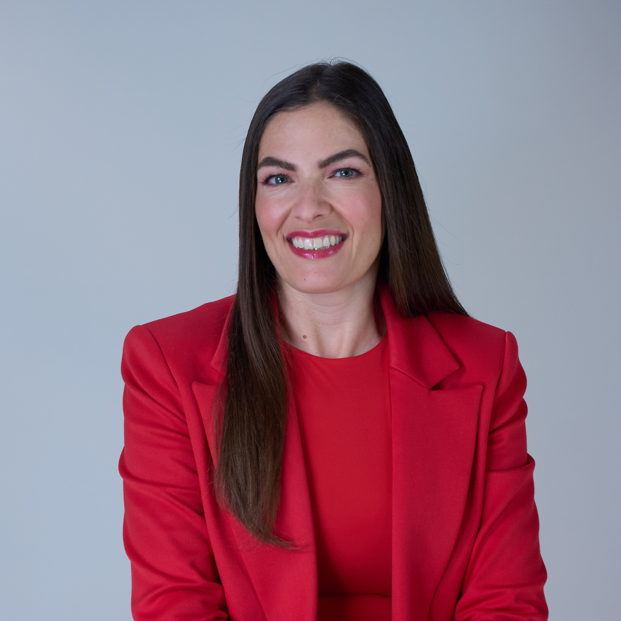 Smiling woman with long dark hair wearing a red blazer and top against a plain light background.