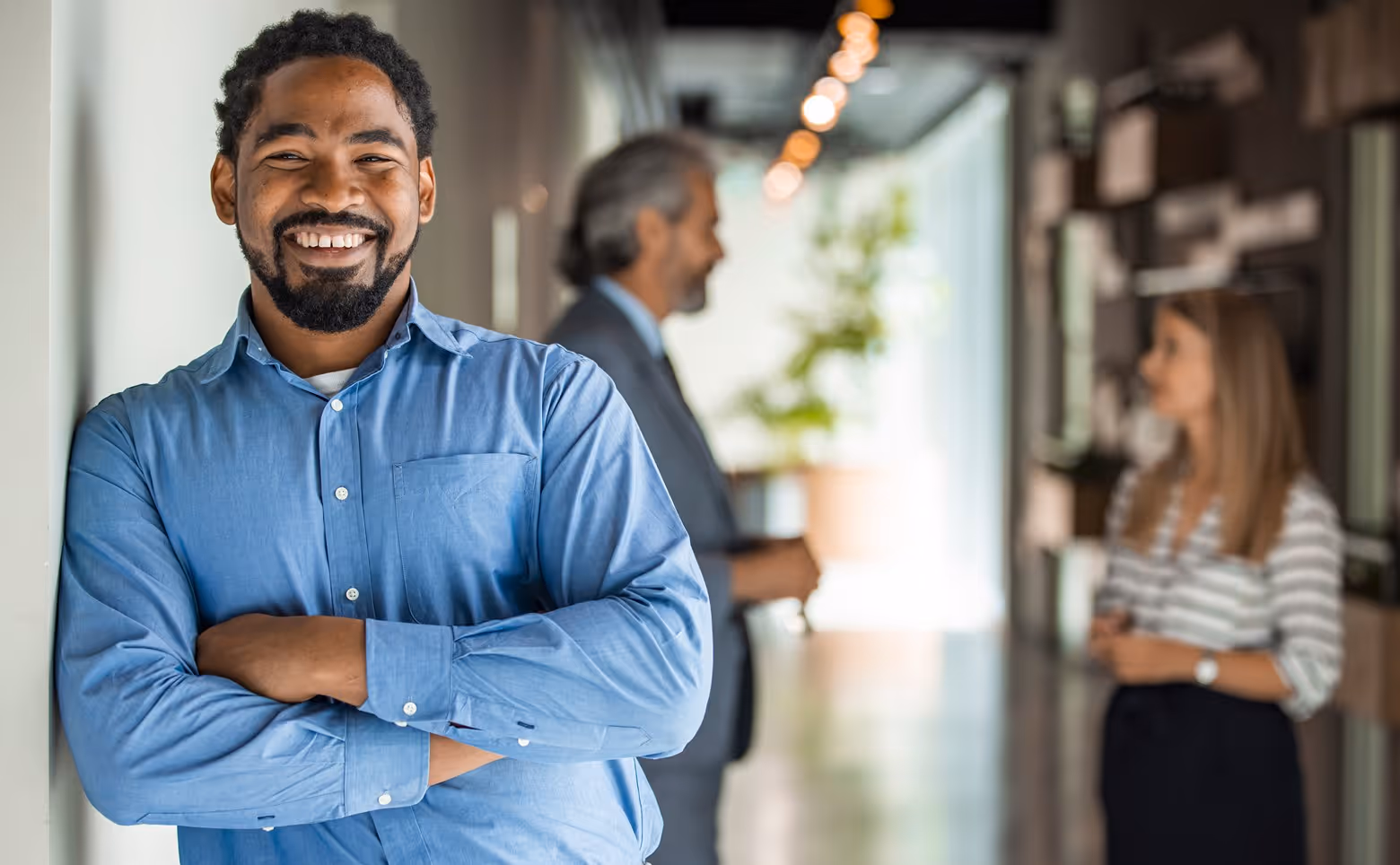 Smiling man in blue shirt standing with crossed arms in office corridor with two colleagues conversing in the background.