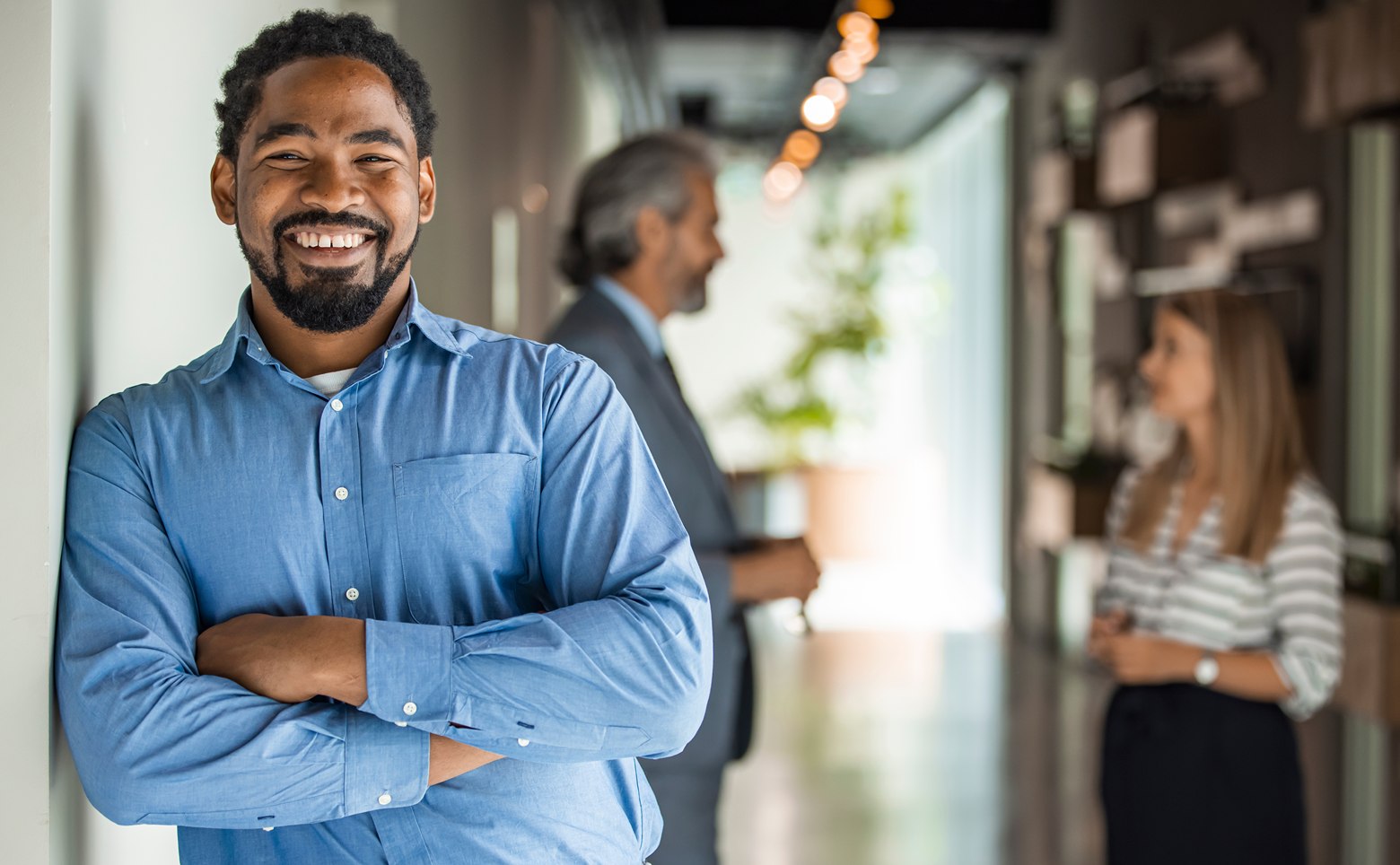 Smiling man in blue shirt standing with crossed arms in office corridor with two colleagues conversing in the background.