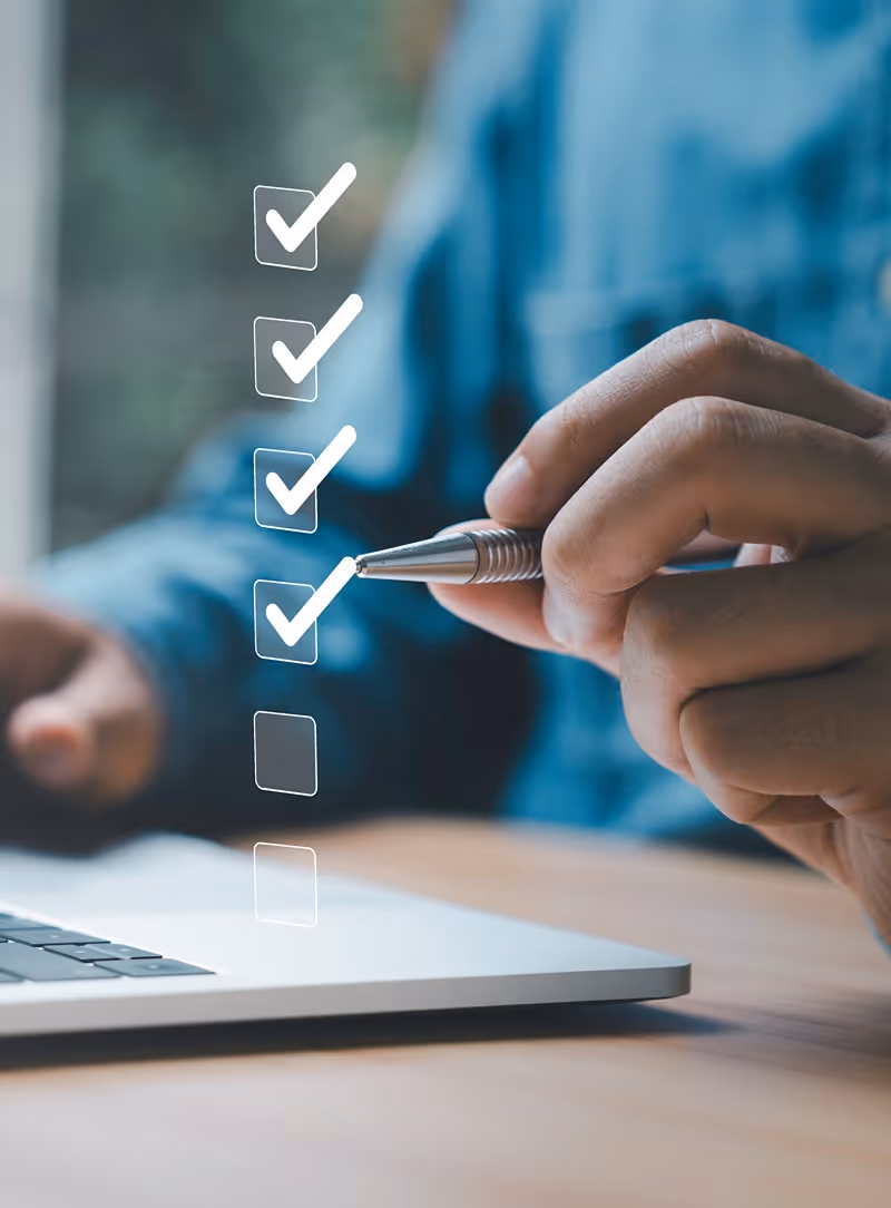 Person holding a pen near a laptop with a digital checklist showing four checked boxes and one unchecked box.