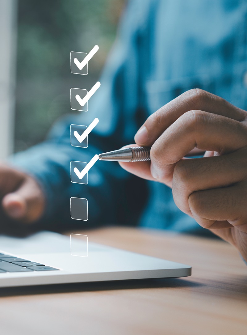Person holding a pen near a laptop with a digital checklist showing four checked boxes and one unchecked box.
