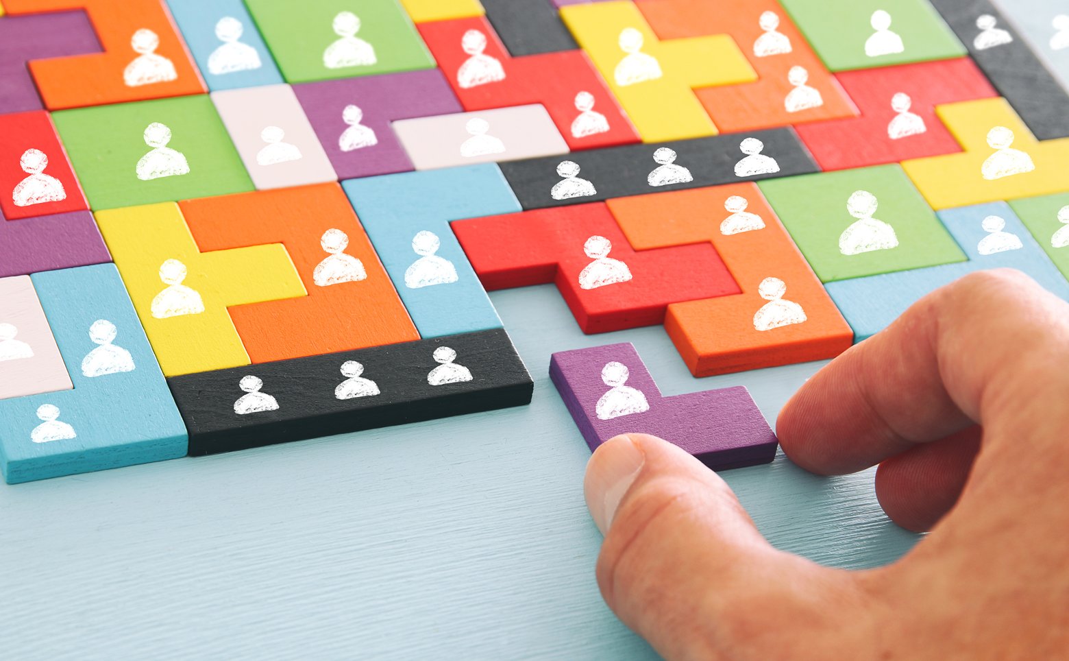 Hand placing a purple L-shaped puzzle piece among colorful interlocking blocks with white person icons on them.