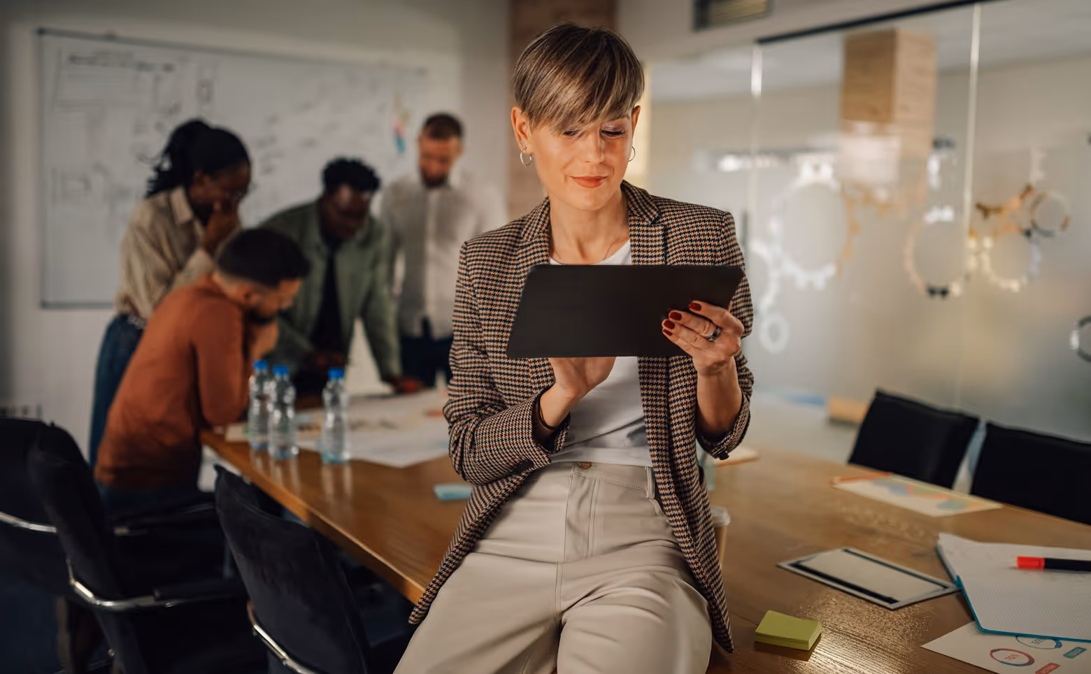 A woman sitting on a conference table using a tablet while four colleagues discuss documents in the background.