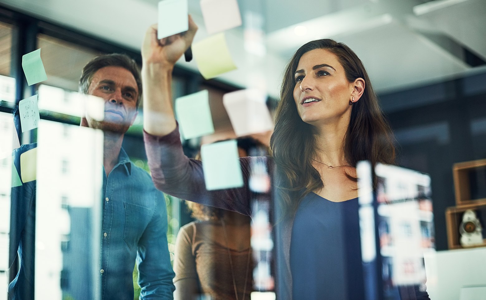 Two colleagues brainstorming and placing sticky notes on a glass wall in a modern office.