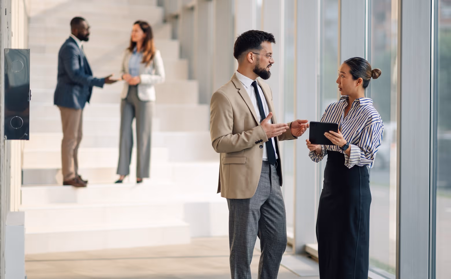 Two pairs of business colleagues engaged in conversations near large windows and a staircase in a modern office.