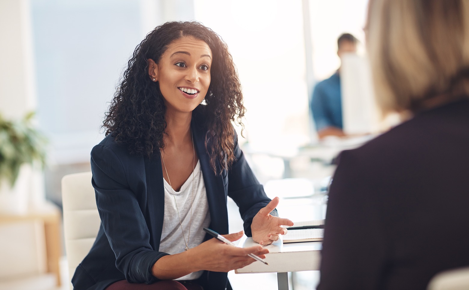 Smiling woman wearing a blazer talking and gesturing during a meeting in a bright office.