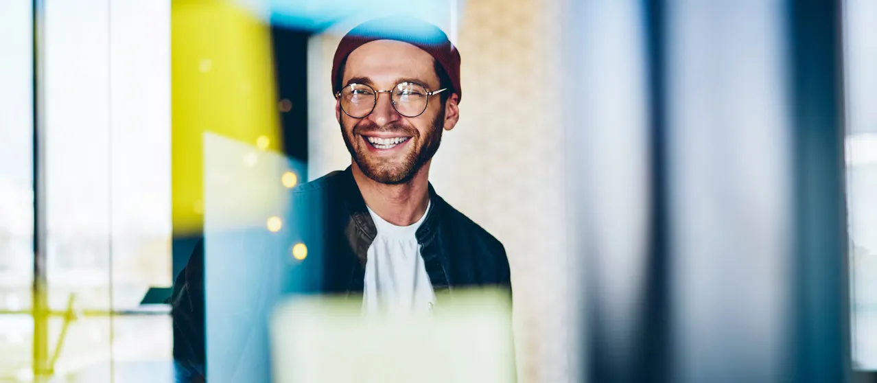 man wearing glasses and beanie smiling through a glass window