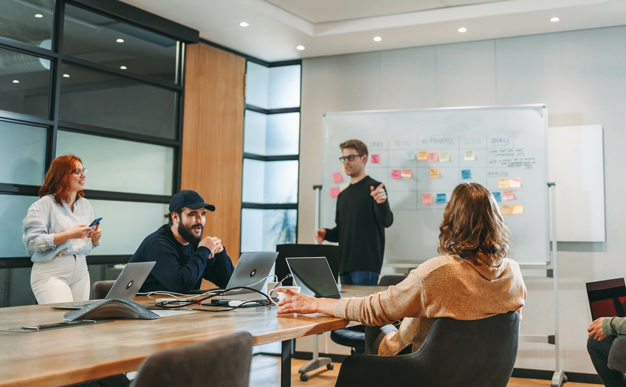 Smiling man in blue shirt standing with crossed arms in office corridor with two colleagues conversing in the background.