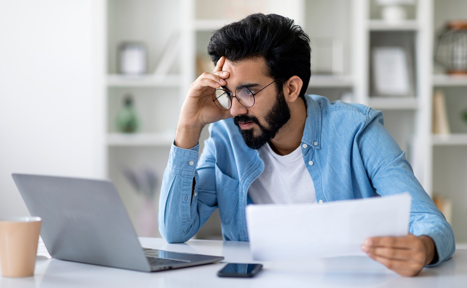 Stressed Young Man Looking At Laptop Screen And Holding Documents