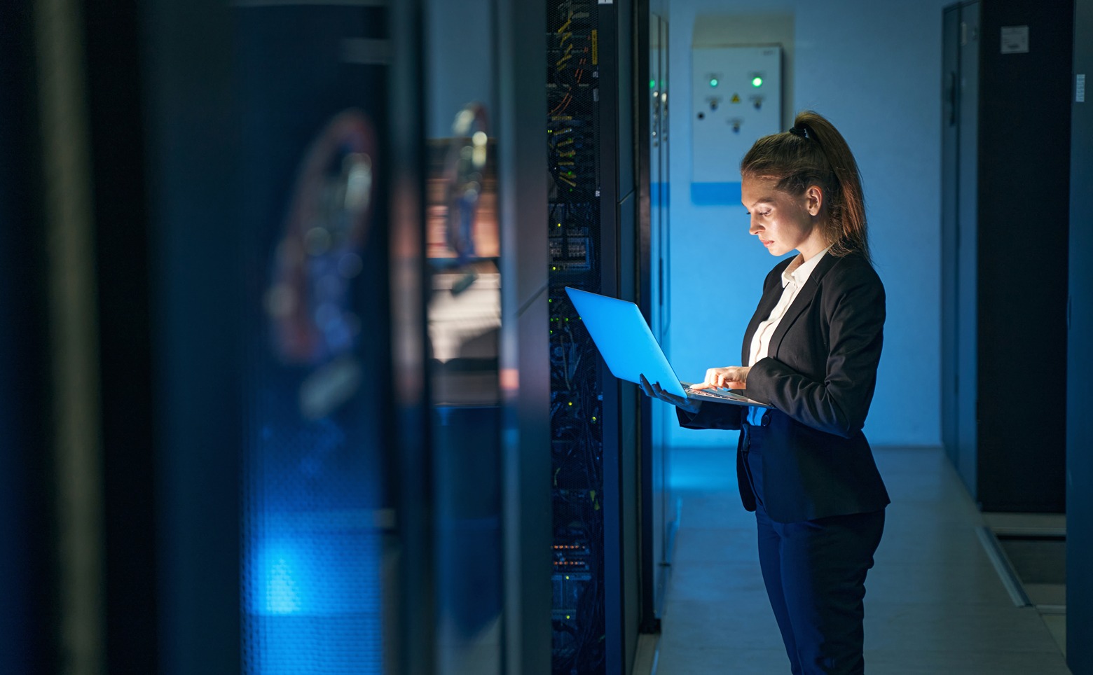 A businesswoman works on a laptop in a dimly lit data center aisle, illuminated by the glow of the screen and server equipment.