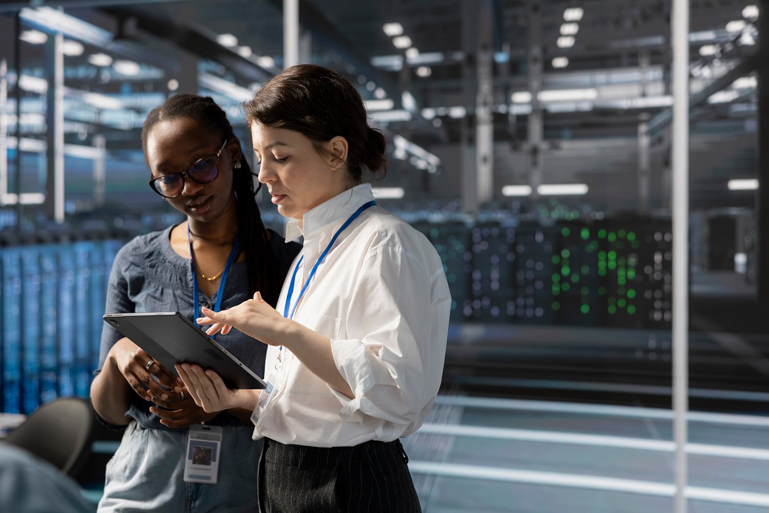 Two women review data on a tablet in a data center, with server racks and status lights visible in the background.