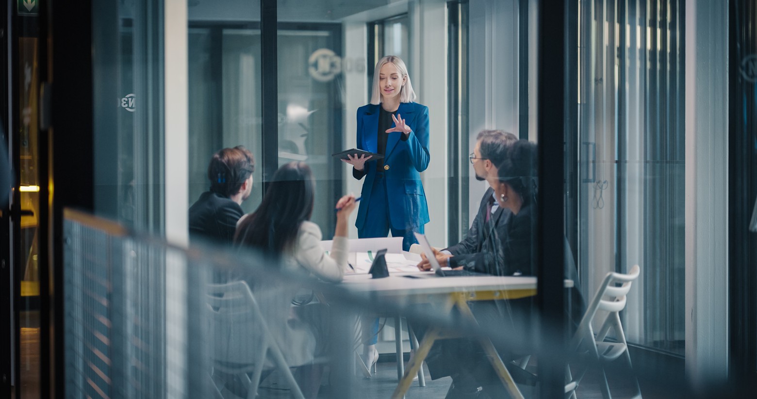 Woman presenting to colleagues around a conference table in a glass-walled meeting room