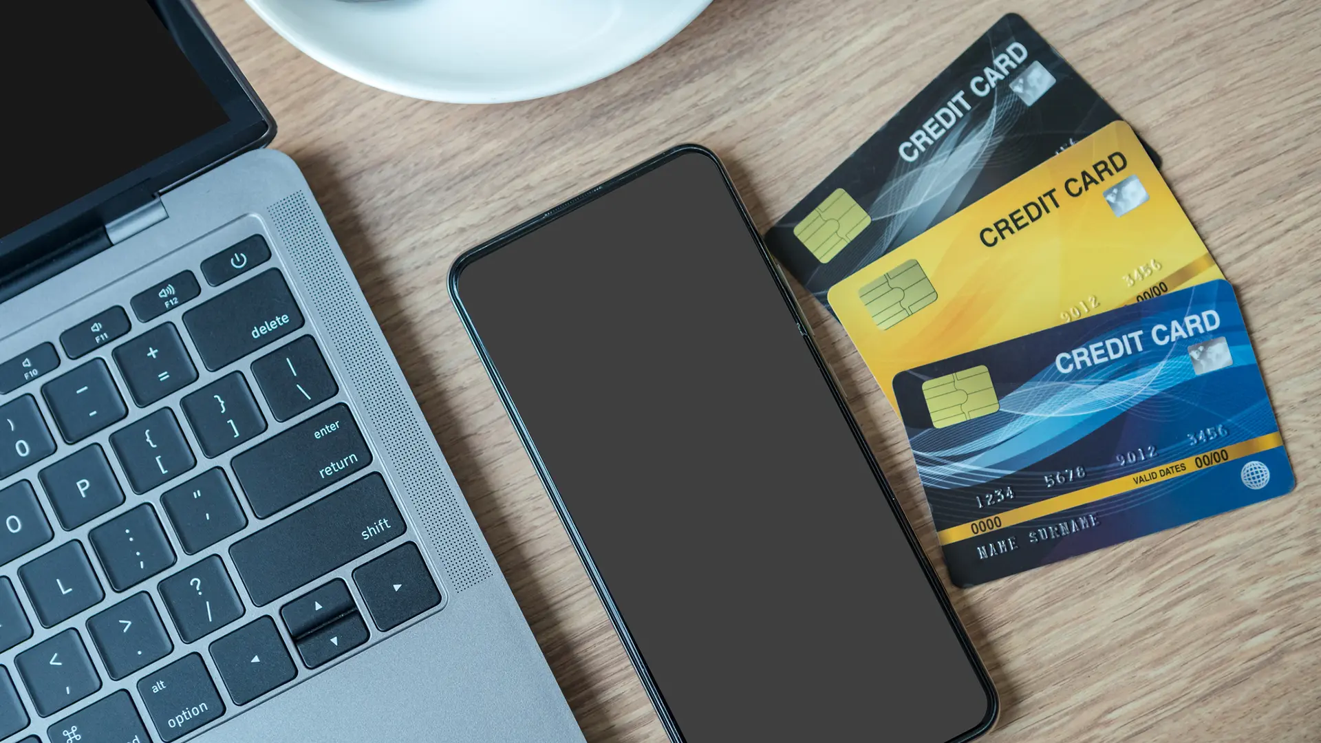 Top-down view of a wooden desk with an open laptop keyboard on the left, a smartphone with a blank screen in the center, and three stacked credit cards on the right, suggesting online finance or credit management.
