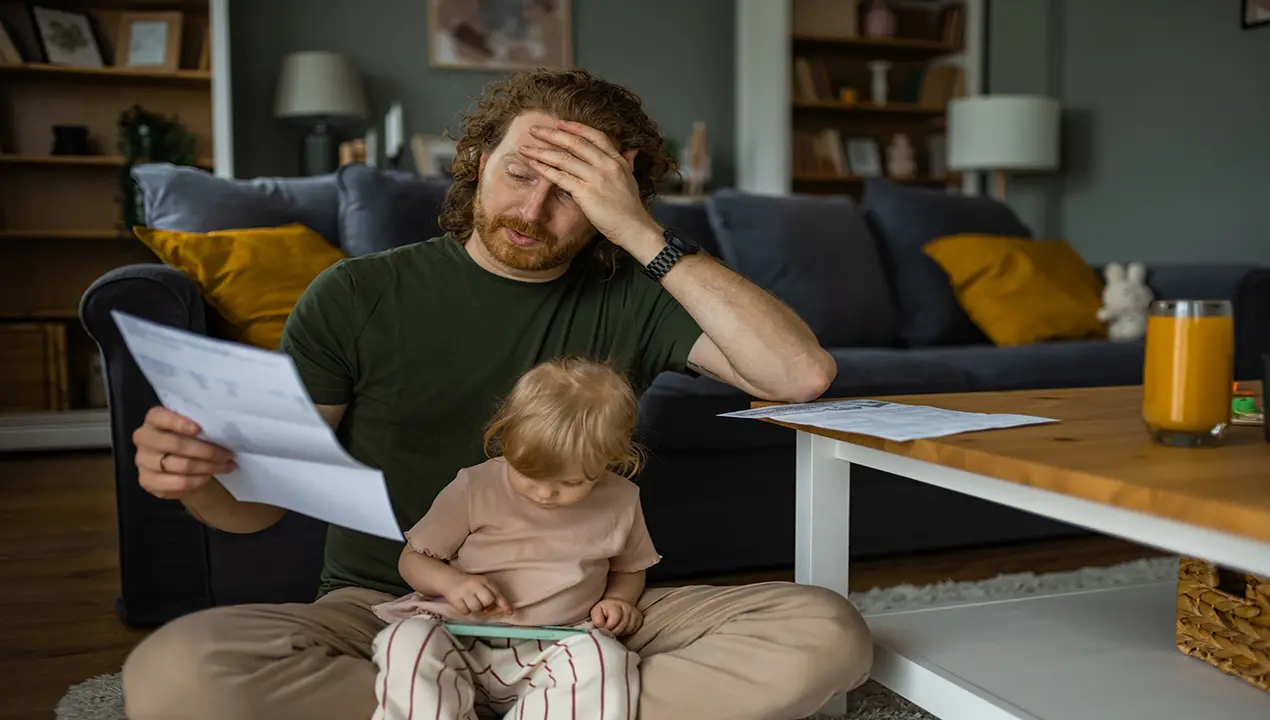 A stressed father holding a bill while sitting with his young child, representing the emotional toll of debt and mental health struggles.