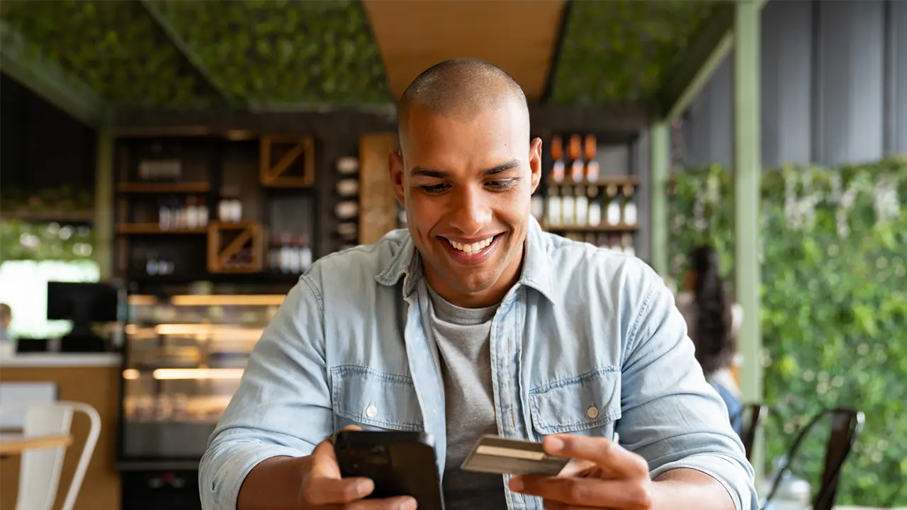 A young man smiling while holding a credit card and phone at a cafe, representing smart credit card habits for building and improving your credit score.