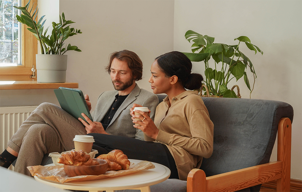 A man in a light gray suit and a woman in a beige sweater sitting on a couch in a cozy café with green plants. They are drinking coffee while looking at a tablet, with a table in front of them holding croissants and cups.