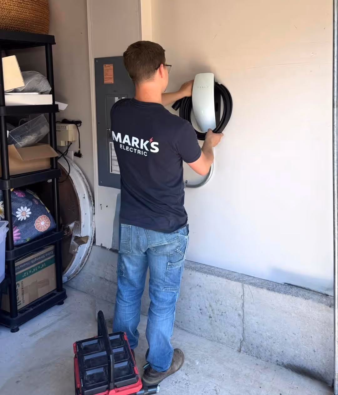 Technician wearing a dark Marks Electric t-shirt installing a Tesla wall charging unit indoors.