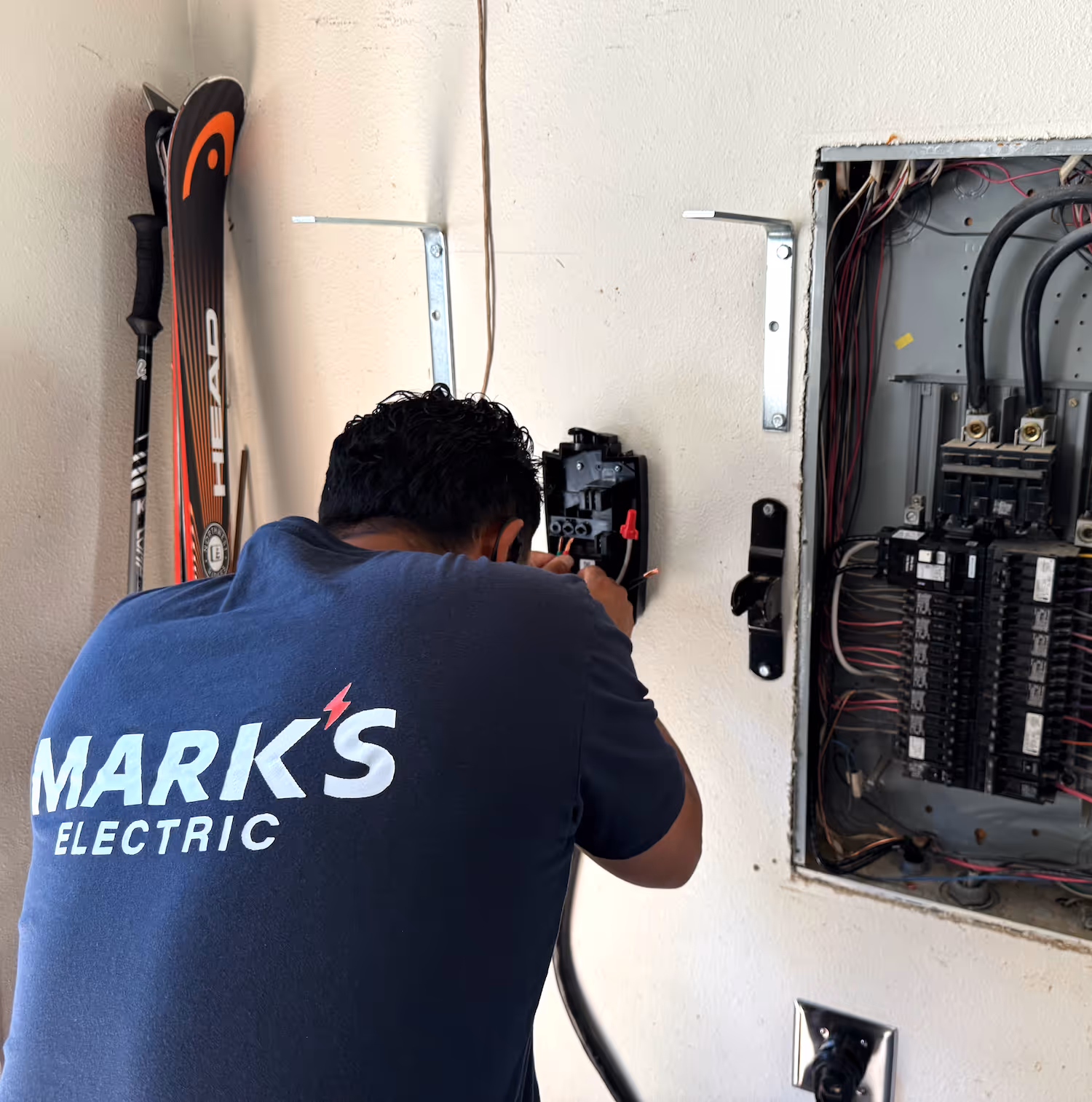 Electrician wearing a Mark's Electric shirt working on wall wiring next to an open electrical panel.