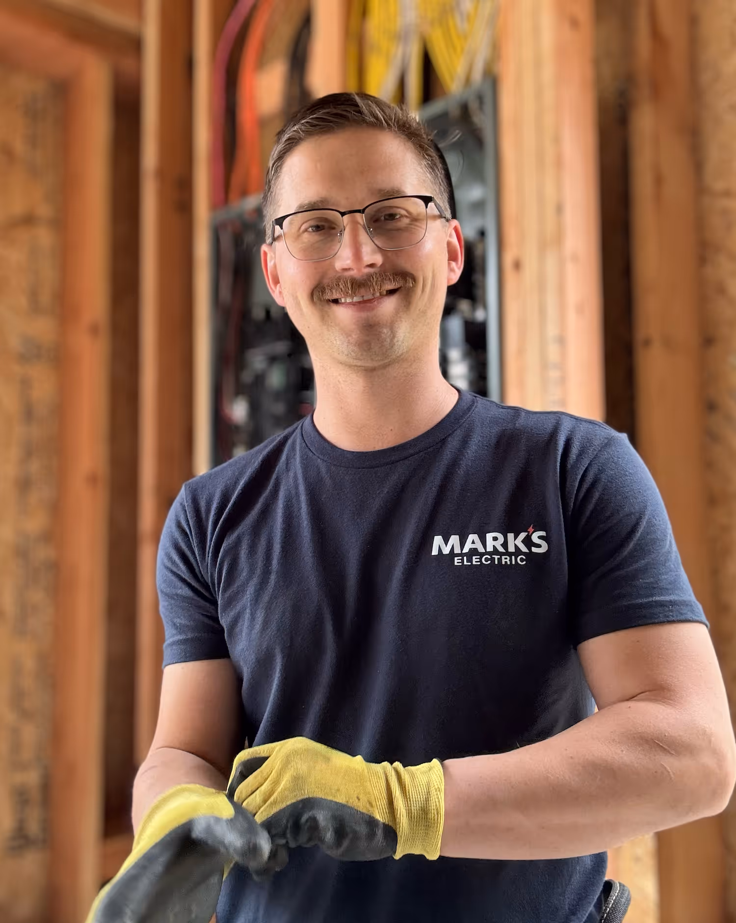 Smiling electrician wearing glasses and yellow gloves, standing inside a wooden framed building with electrical wiring.