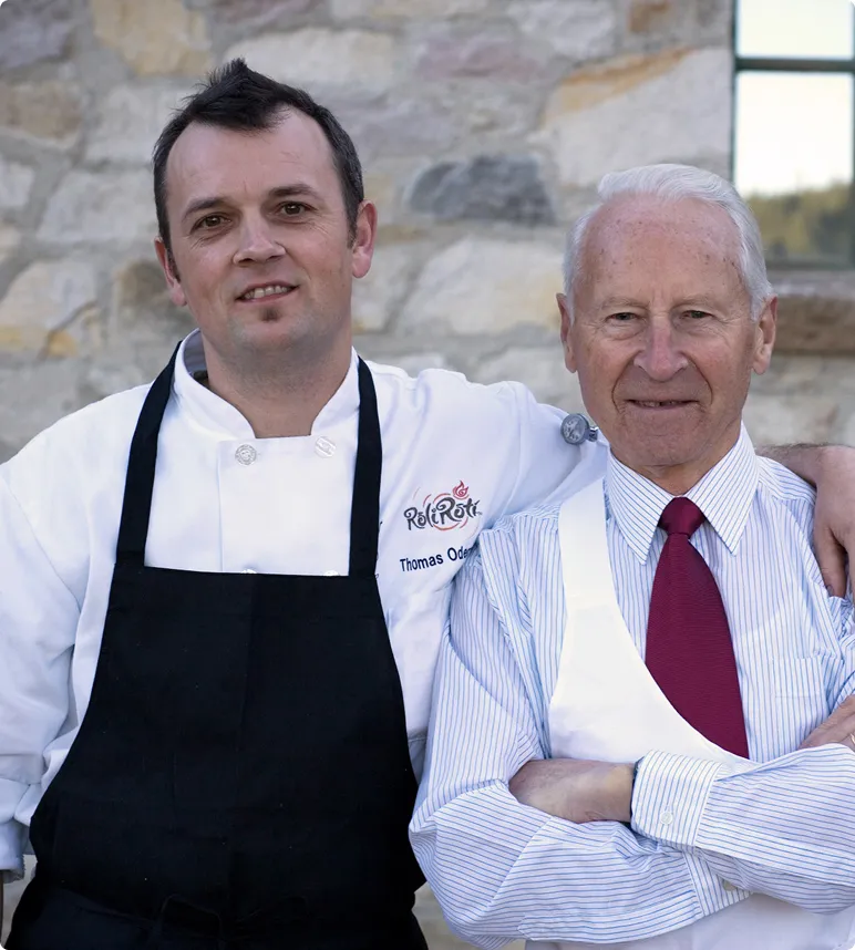 Photograph of young man at left, Thomas Odermat, founder, next to an older man at right, his father Otto. Both men wearing aprons and standing in front of a stone wall.