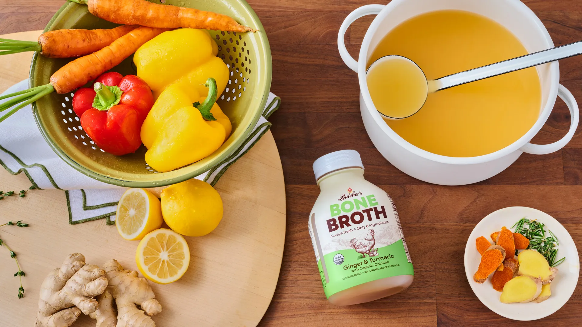 Overhead photograph of a kitchen countertop with a strainer of colorful vegetables and a bottle of Butcher's Bone Broth and stock pot