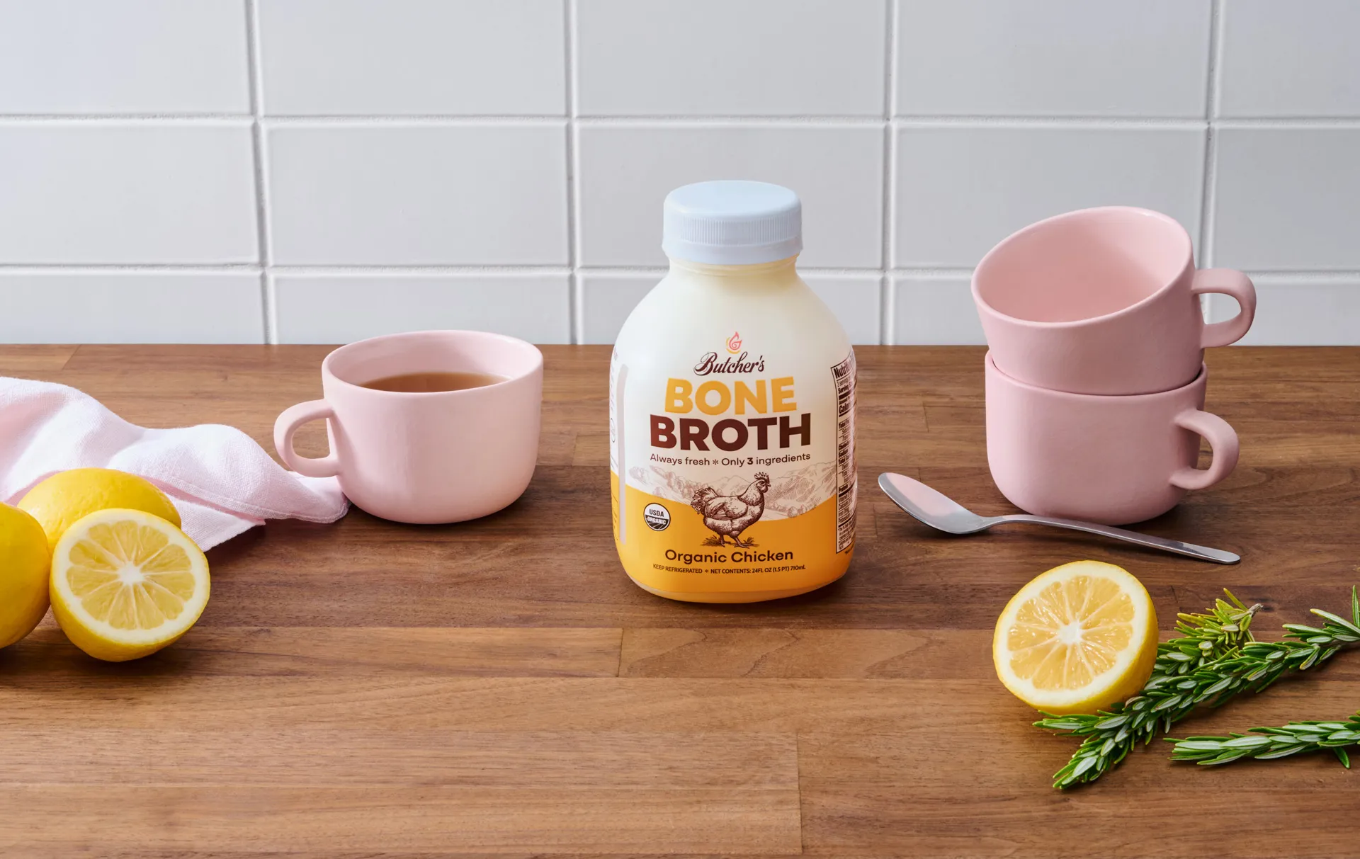 Bottle of Butcher's Bone Broth chicken flavor on kitchen counter with pink mugs and lemons