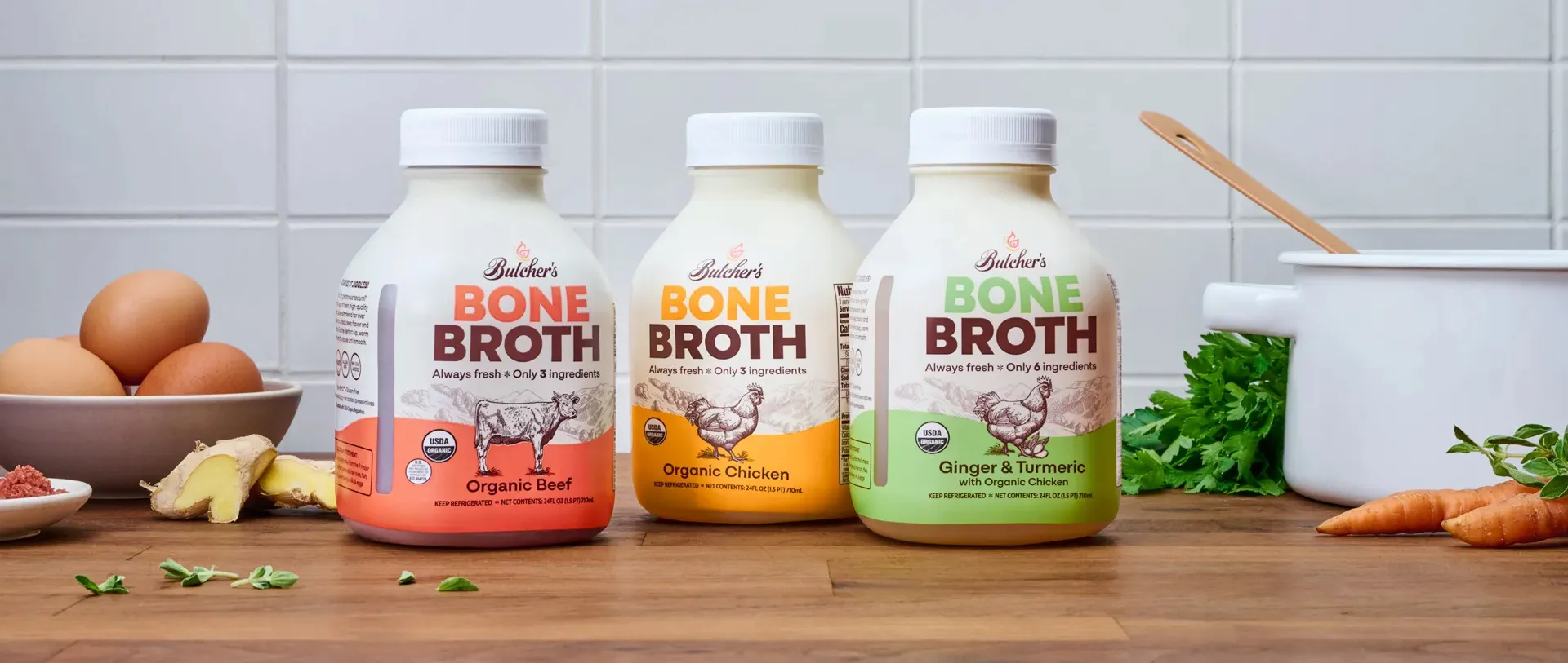 Trio of Butcher's Bone Broth bottles (red, yellow, green) sitting on kitchen counter with food and stock pot