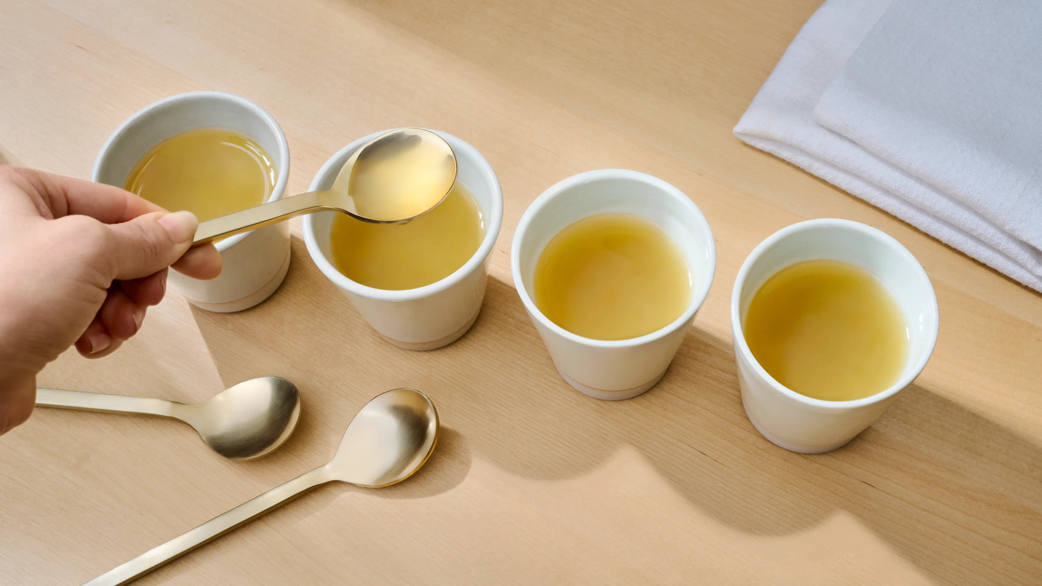 Four cups of broth with hand holding a brass colored tasting spoon