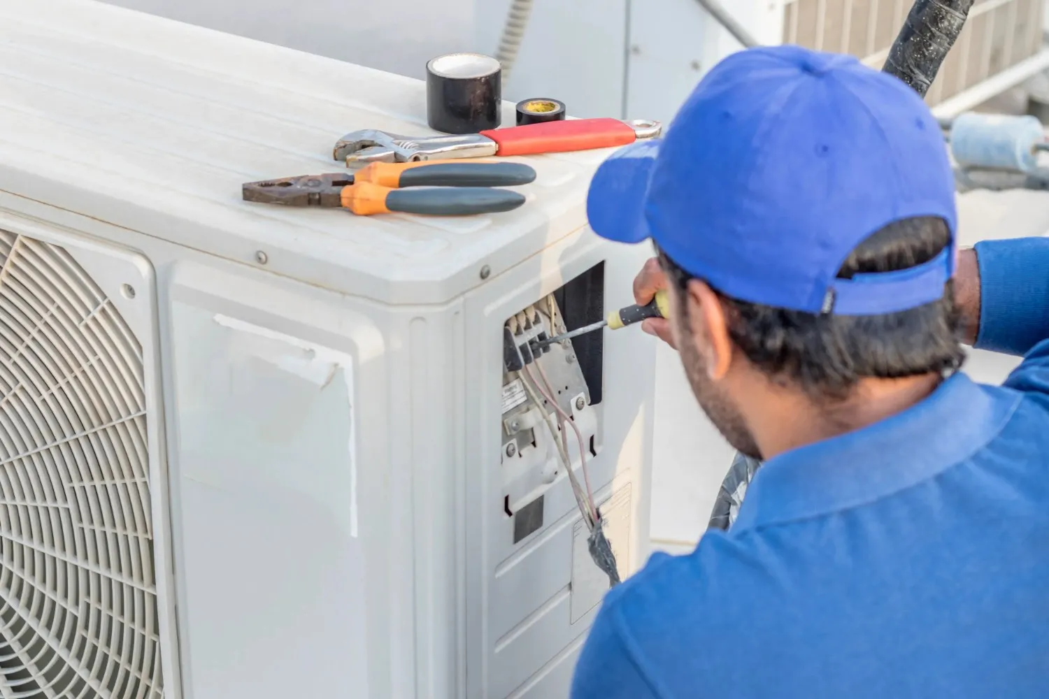 A close-up shows hands working with tools on the top of a white outdoor air conditioning unit