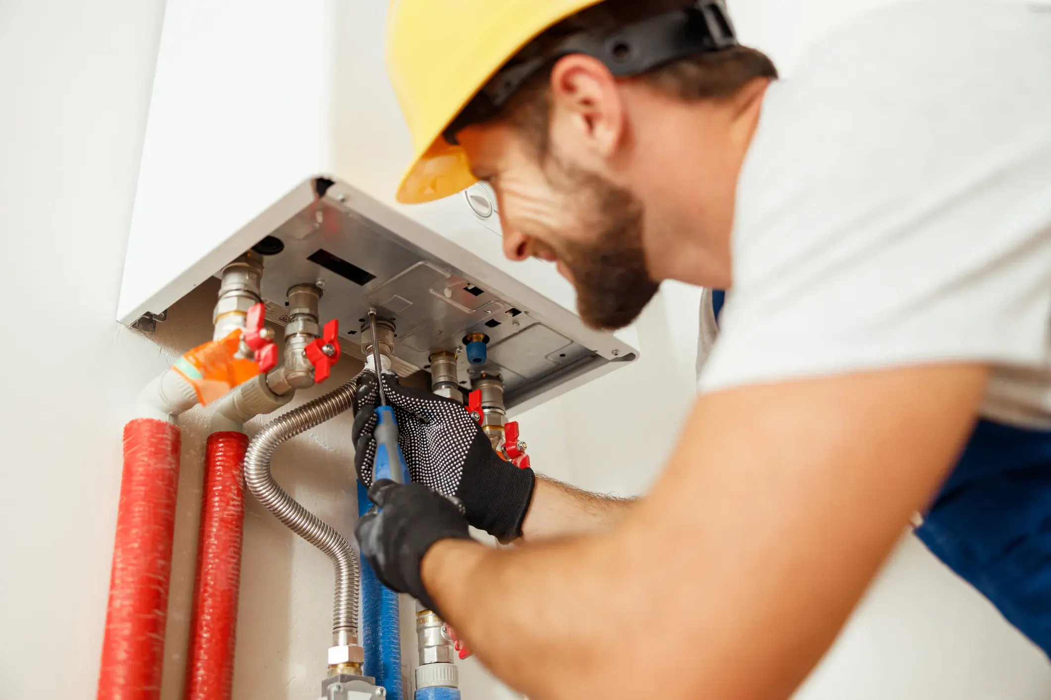 A male plumber wearing a yellow hard hat and safety glasses is using a wrench to work on the pipes of a wall-mounted tankless water heater.
