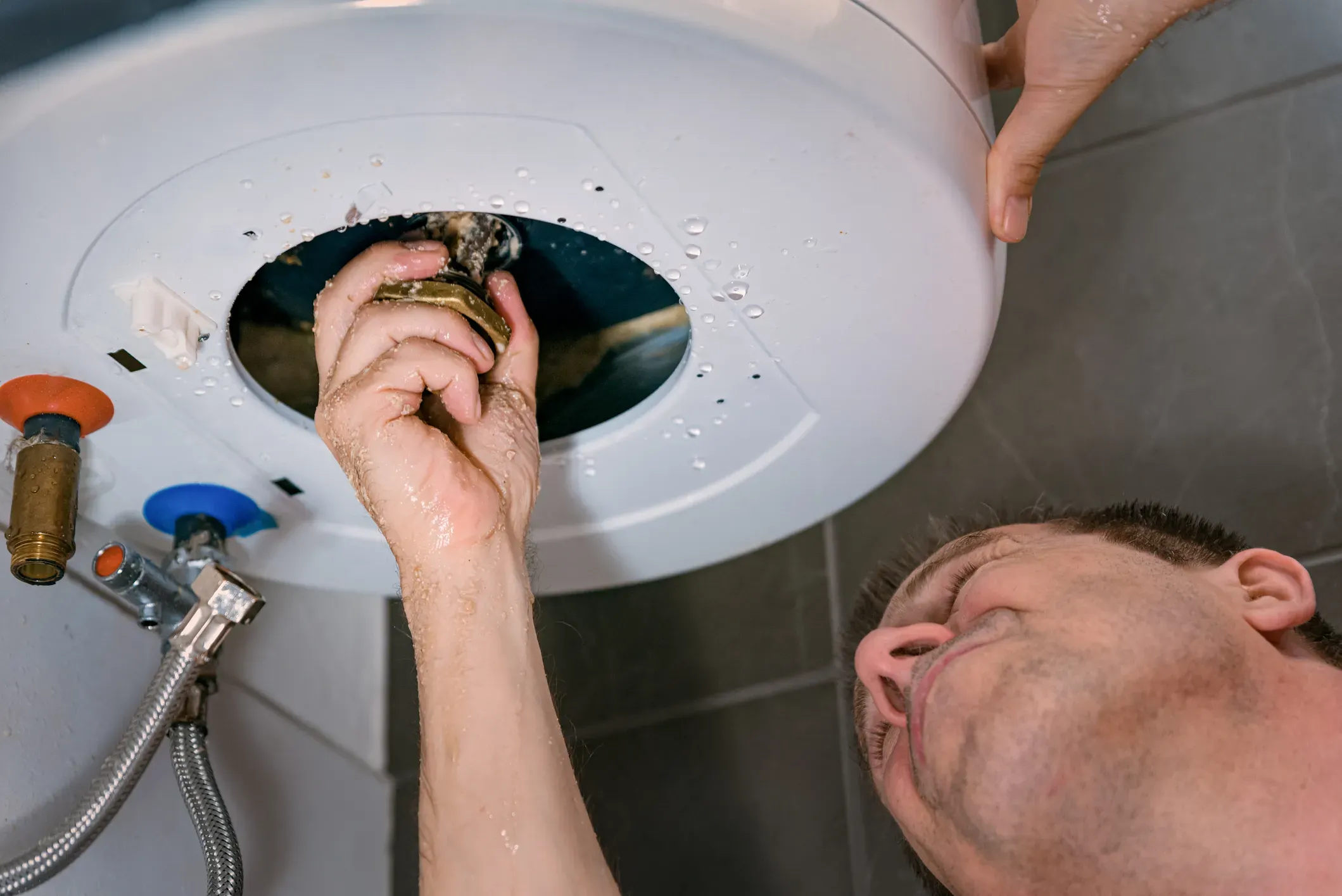 A plumber is shown from a low angle, reaching up to remove a component from the bottom of a water heater with water dripping from it.