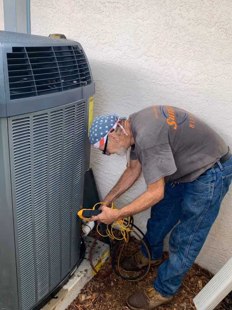 Technician in patriotic cap repairs air conditioning unit outside building