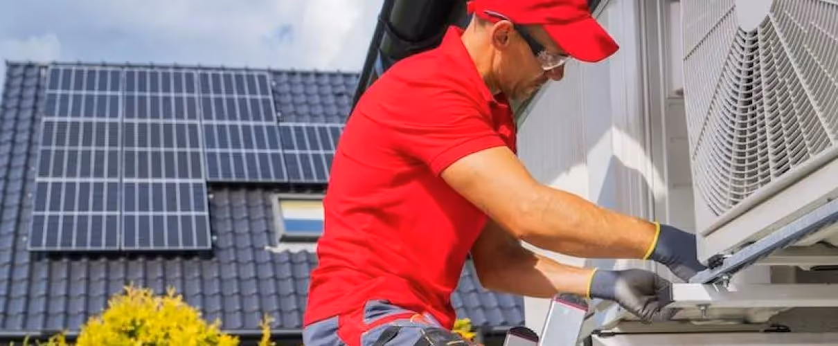 Technician in red uniform installing or repairing air conditioning unit near solar panels