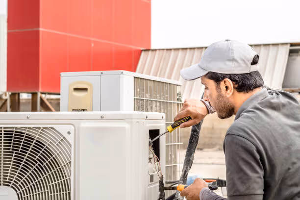 Technician in cap repairing air conditioning unit against red building
