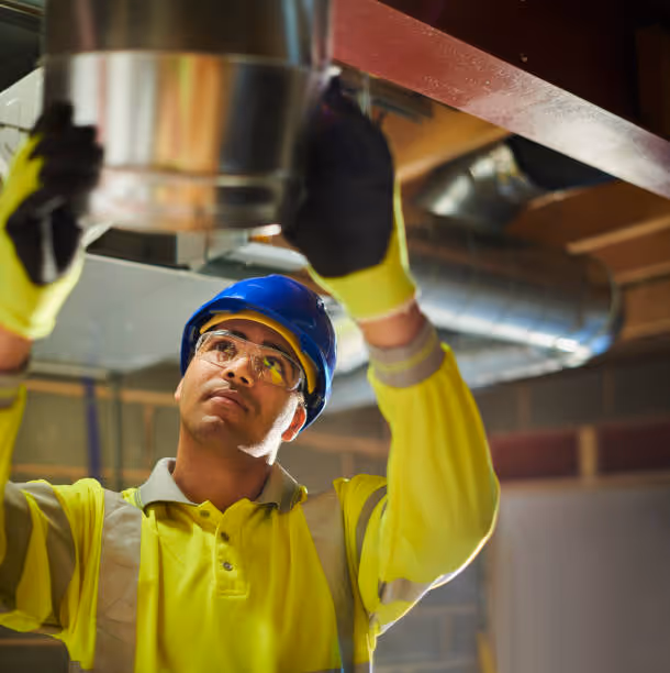 Worker in safety gear inspects overhead ductwork in industrial setting