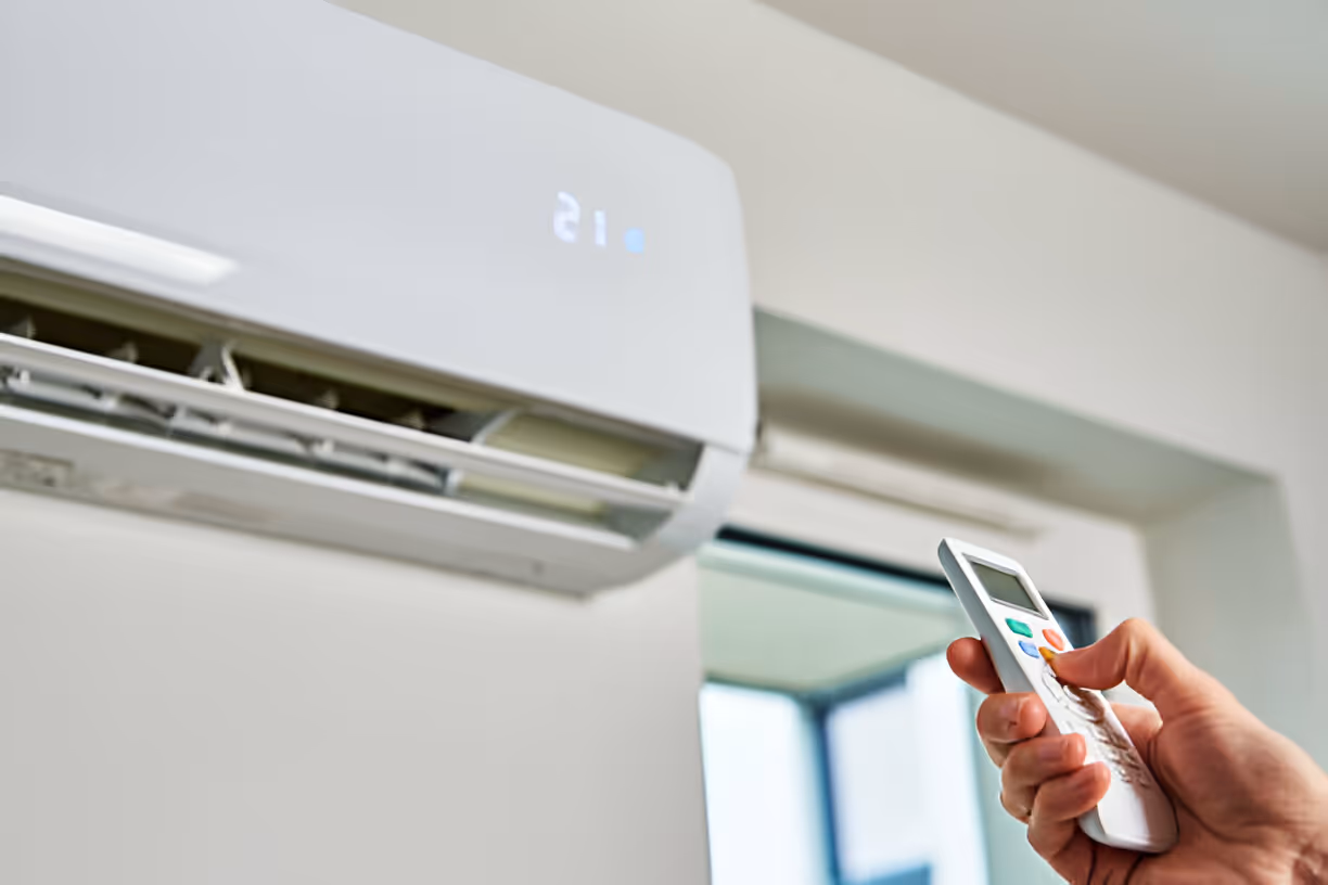 Hand holding remote control aimed at wall-mounted air conditioning unit