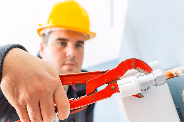 Plumber in hard hat using red pipe wrench on white plumbing connection