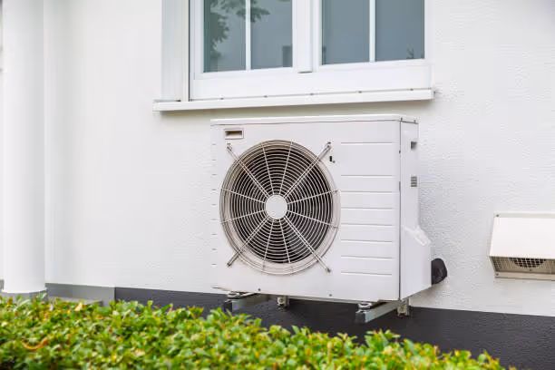 White air conditioning unit mounted on exterior wall near green bushes
