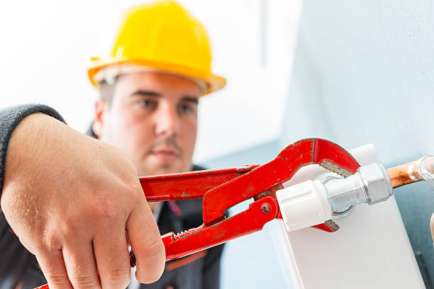 Plumber in yellow hard hat using red pipe wrench on white pipe