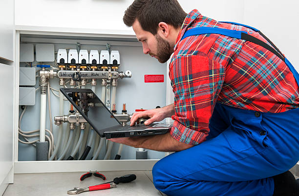 Technician in plaid shirt checks heating system manifold with laptop