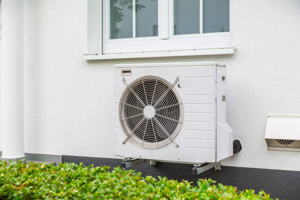 White air conditioning unit mounted on exterior wall near green bushes