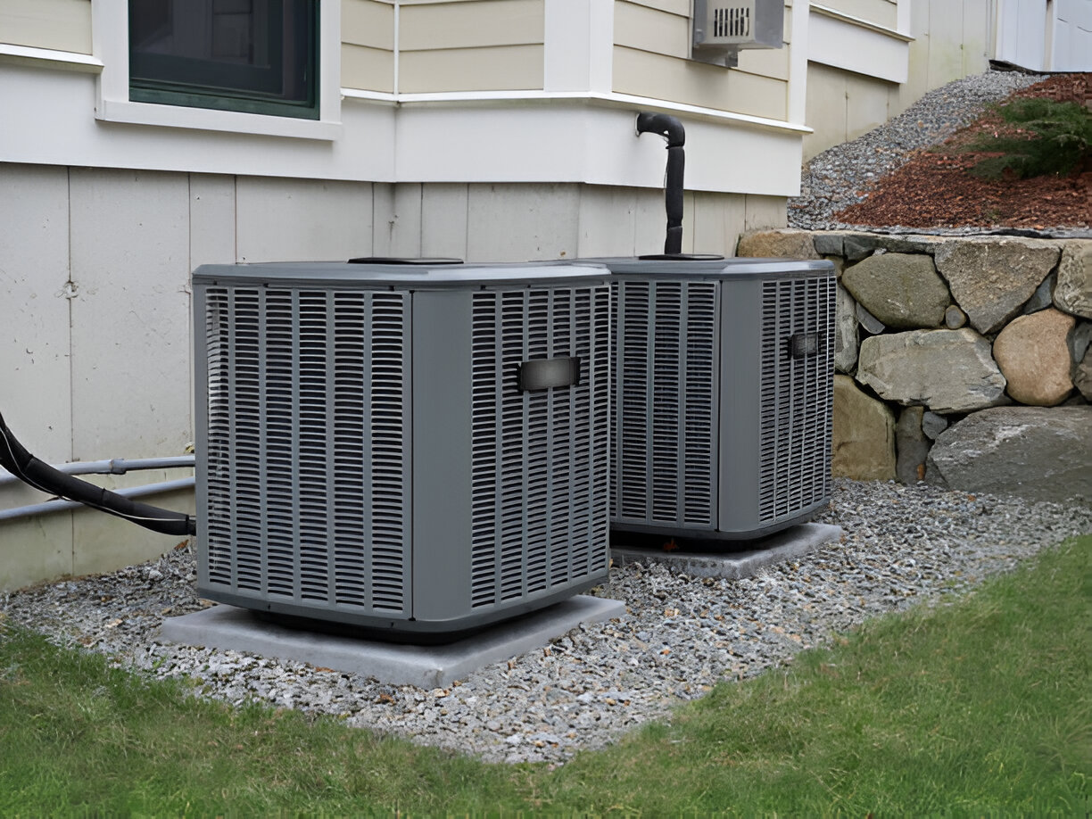 Two gray air conditioning units outside a house near a stone wall