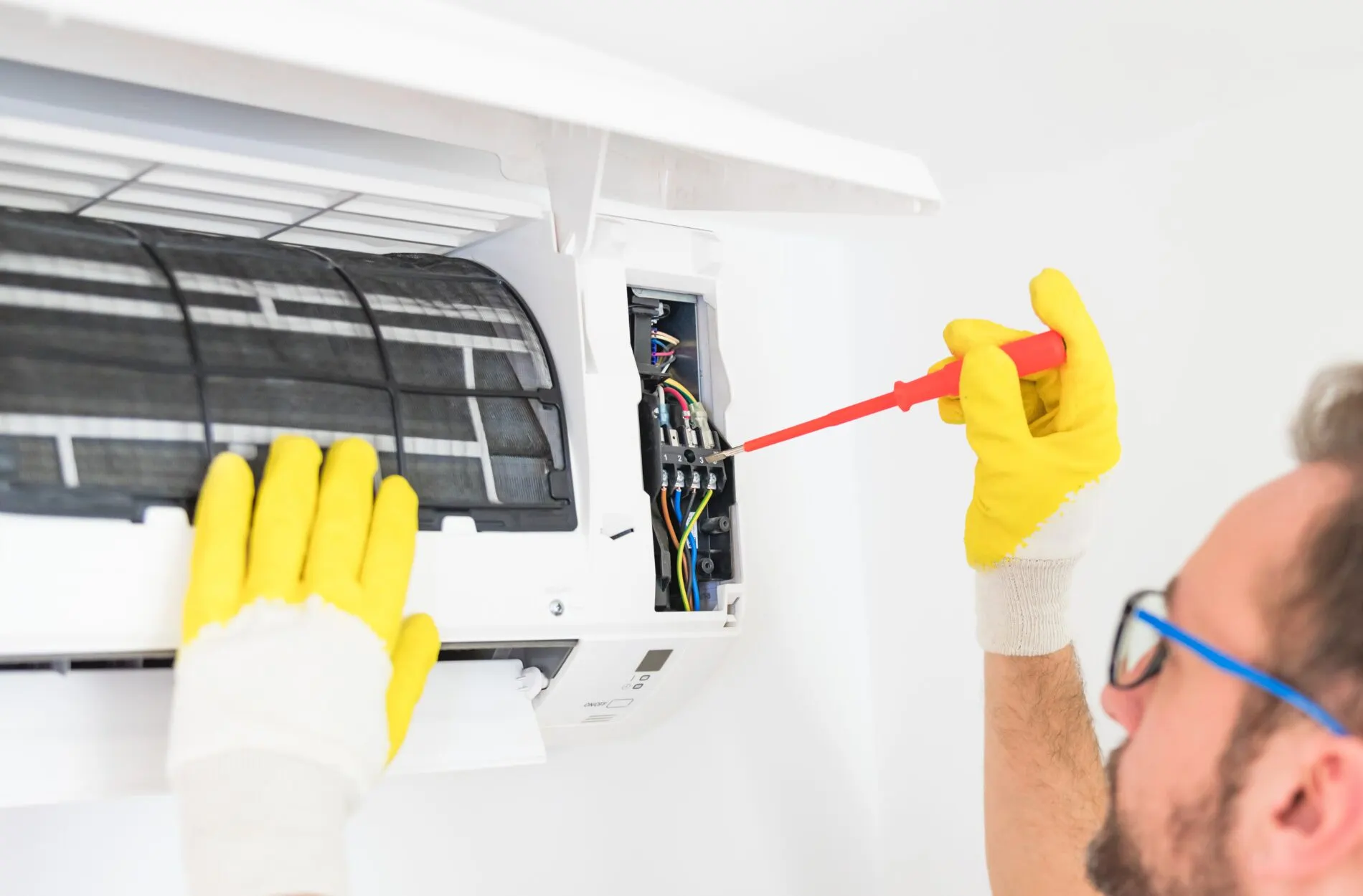 A technician wearing yellow gloves and safety glasses is performing maintenance on an indoor mini-split air conditioner unit.