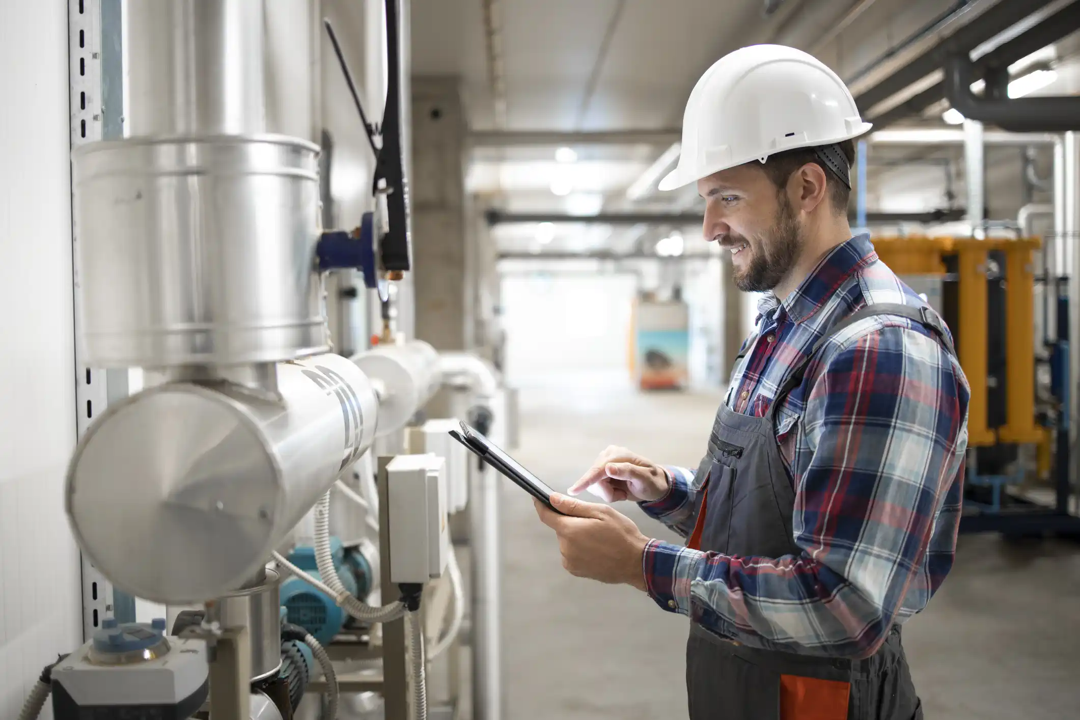A smiling male engineer or technician wearing a white hard hat, a plaid shirt, and gray overalls is standing in a commercial or industrial utility room. He is looking down and interacting with a black digital tablet he holds in his hands. Insulated metal piping and various controls are visible on the wall to his left.