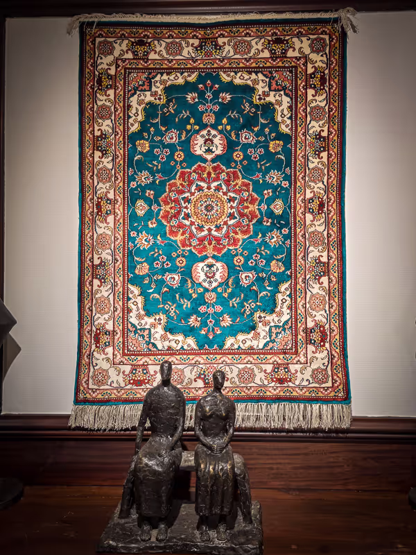 Een kleurrijk, gedetailleerd tapijt hangt aan de muur achter twee zittende metalen sculpturen.